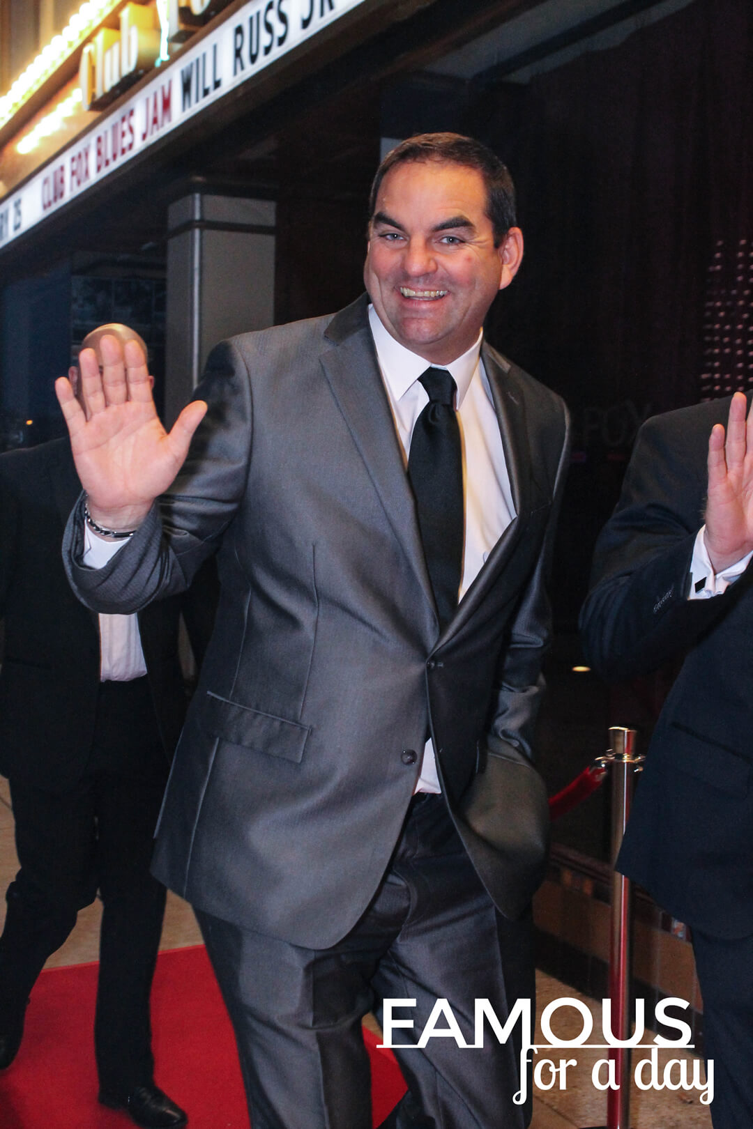 Man in a suit waving and smiling while walking along a red carpet outside a venue